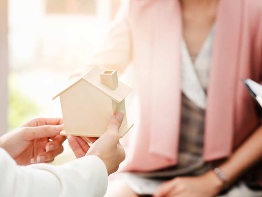 Hands holding up a wooden model of a house, showing it to a woman sitting across