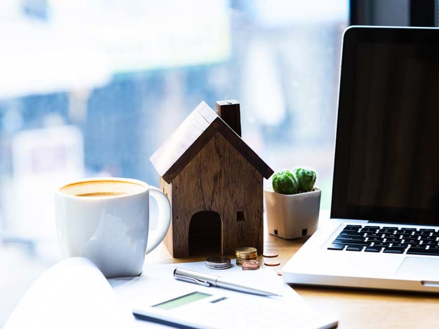 Small wooden house on a desk next to a laptop and a cup of coffee